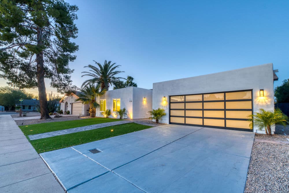 Modern white house with large windows, palm trees, and a spacious driveway at dusk.